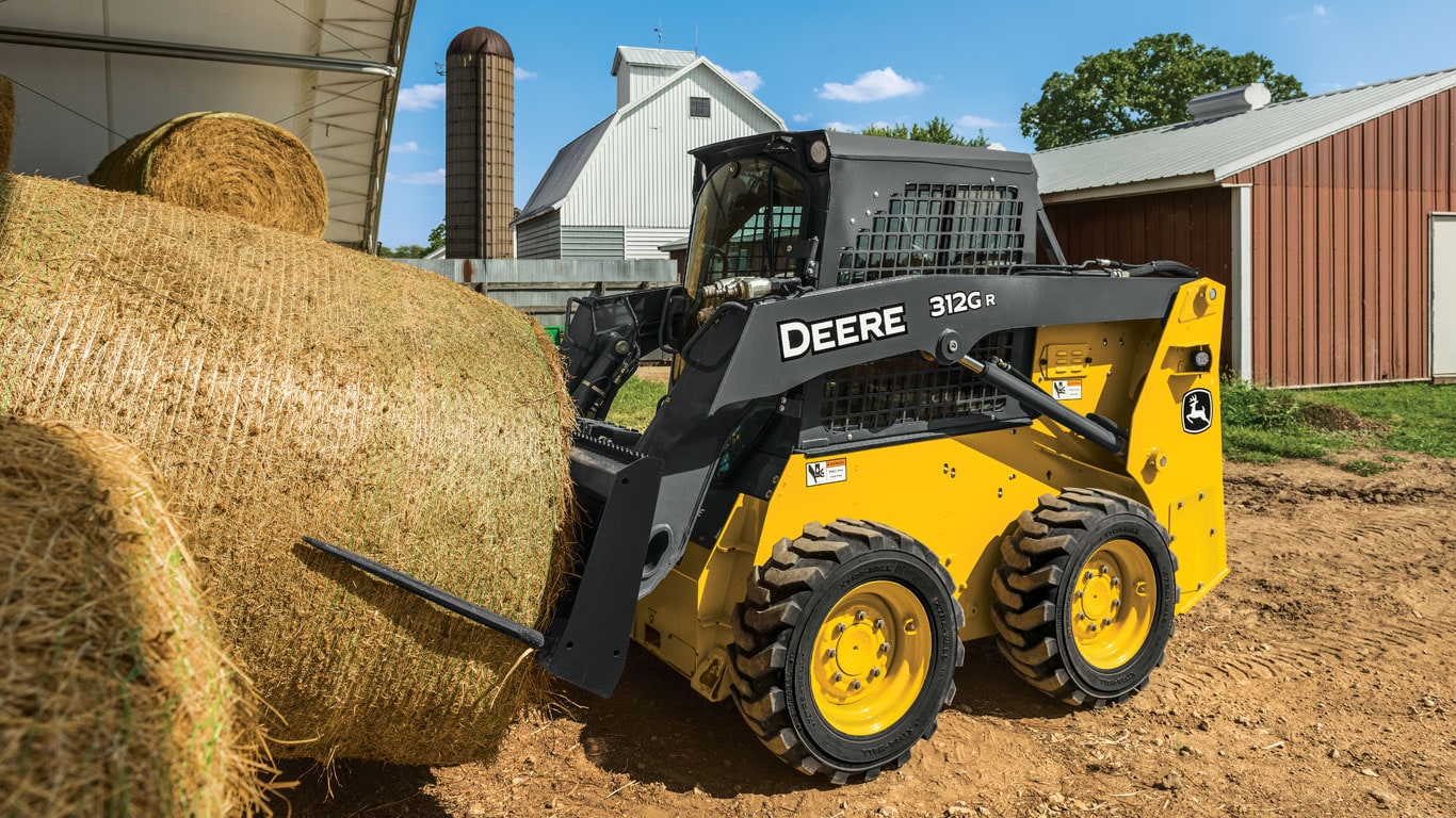 Une chargeuse à direction différentielle John Deere équipée de l’accessoire pointe à balles déplace une balle.